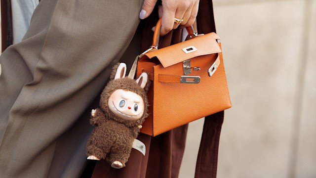 A guest wears brown coat, white blouse, orange Hermes bag with a bag charm brown Labubu monster, grey pants outside the Hermes fashion show during the Womenswear Fall/Winter 2025/2026 as part of Paris Fashion Week on March 08, 2025 in Paris